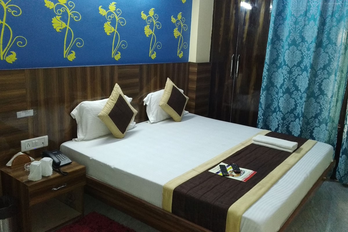 A hotel room with a wooden headboard, a bed with white bedding and two pillows, a nightstand with a phone and tissues, and a blue patterned curtain.