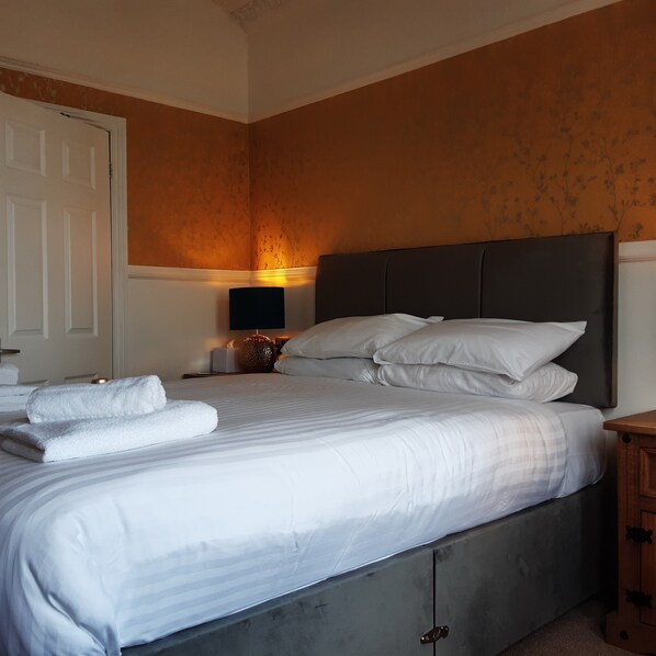 A neatly made bed with white linens and pillows, a bedside lamp, and a wooden nightstand with a small decorative item.
