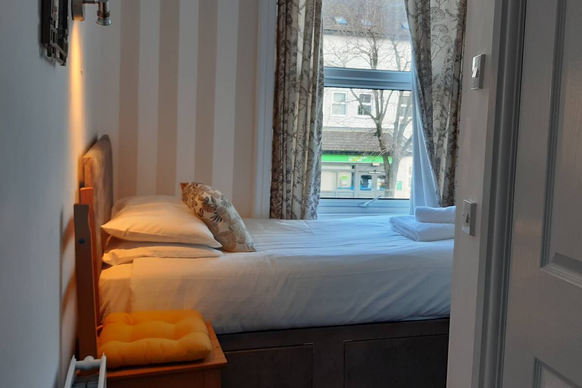 Een slaapkamer met een bed, een raam met gordijnen, een radiator en een klein krukje.