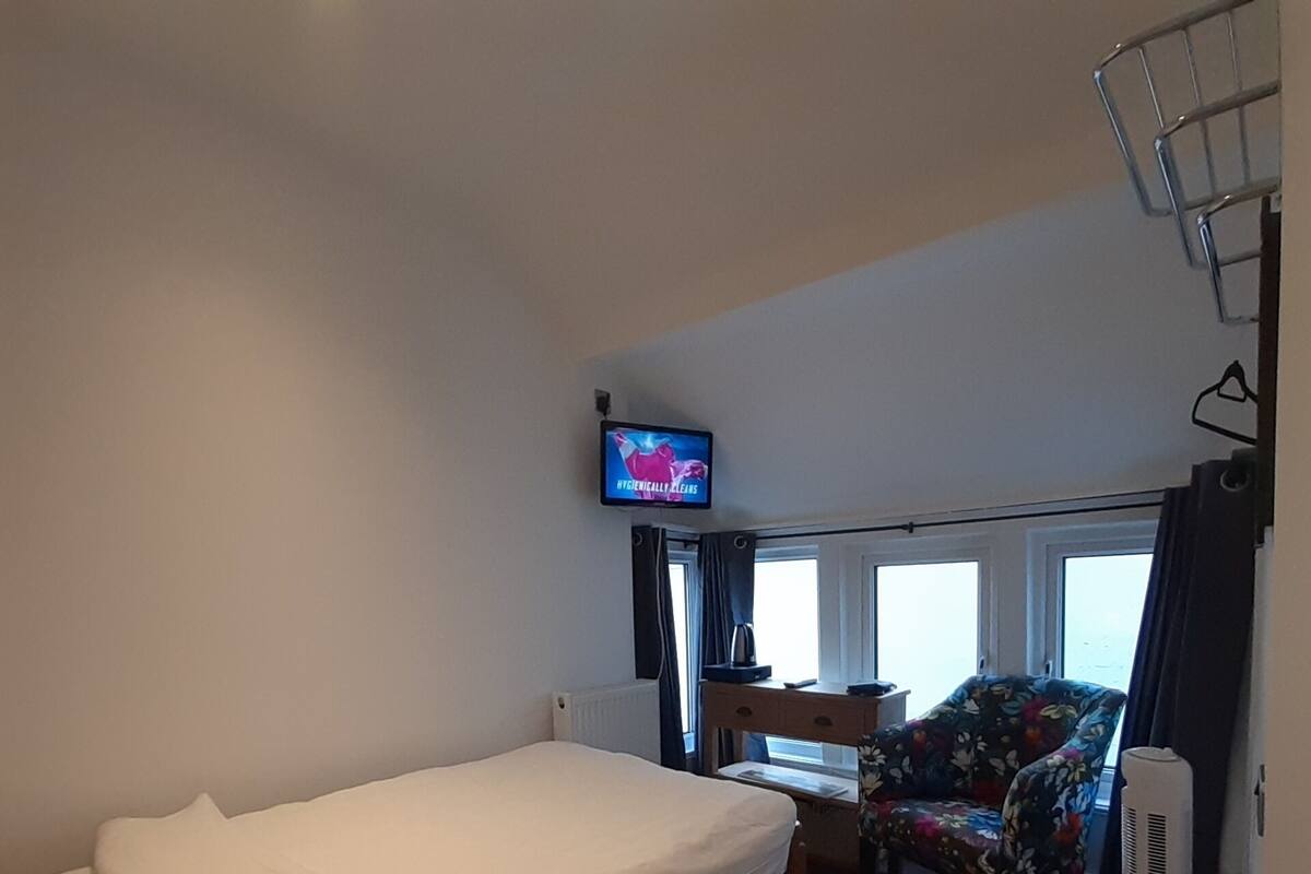 Een hotelkamer met een enkel bed, een bureau met stoel, een aan de muur bevestigde televisie en een raam met gordijnen.