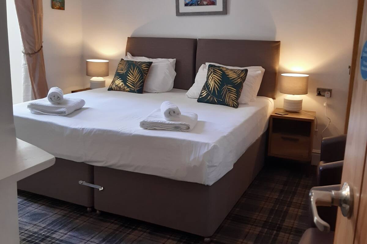Een net opgemaakt bed met witte lakens en twee decoratieve kussens, een nachtkastje met een lamp, en een vloer met een geruit patroon.