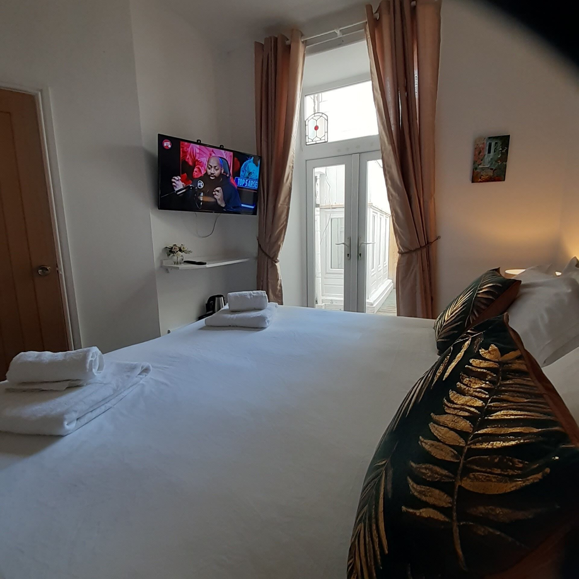 A hotel room with a bed, a TV mounted on the wall, a window with curtains, and a small shelf with a vase.