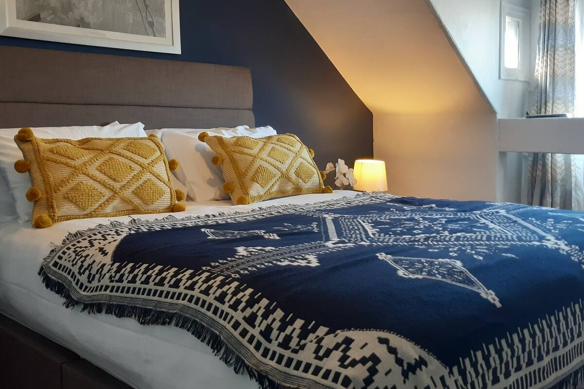 Een slaapkamer met een bed, kussens, een deken, een nachtkastje, een lamp en een ingelijst foto aan de muur.