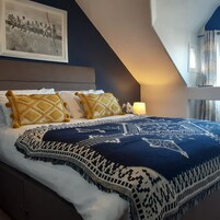 Un dormitorio con una cama, almohadas, una colcha, una mesita de noche, una lámpara y un cuadro en la pared.