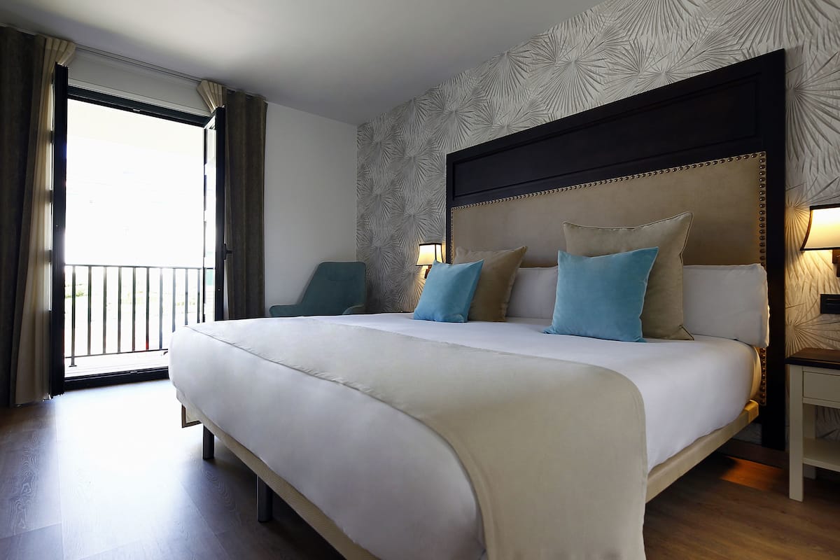 A large bed with a headboard, a nightstand, a chair, and a view of the outside.