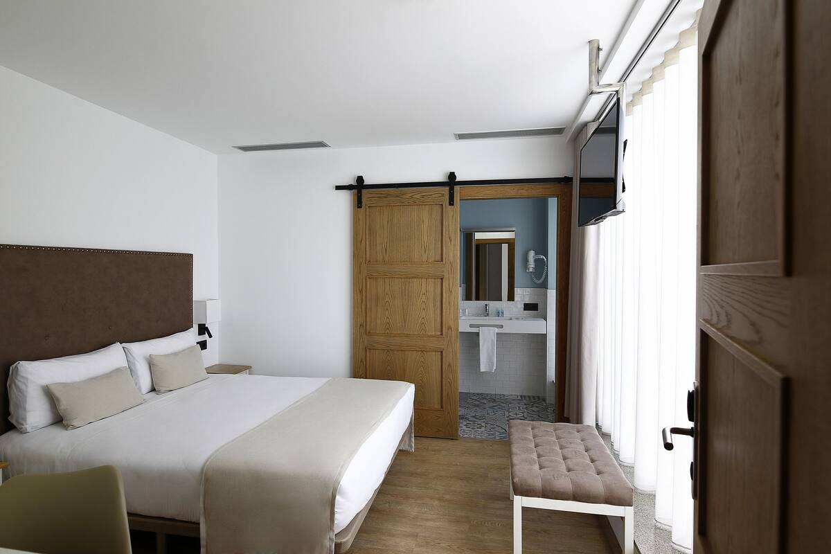 A hotel room with a large bed, a wooden door, a bathroom with a mirror, and a bench.