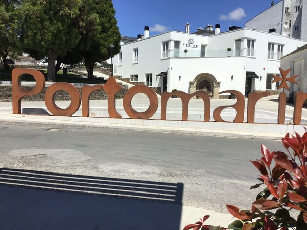 A sign with the name 'Portomarino' in large letters, a white building with balconies, and a tree in the background. - Hotel Vistalegre (Portomarin)