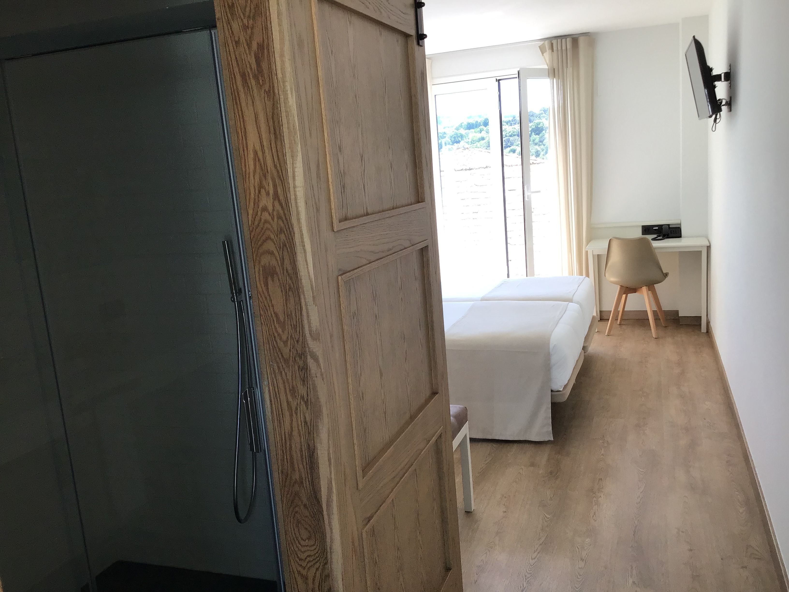 A hotel room with a wooden wardrobe, a bed, a desk with a chair, and a view of the outdoors.