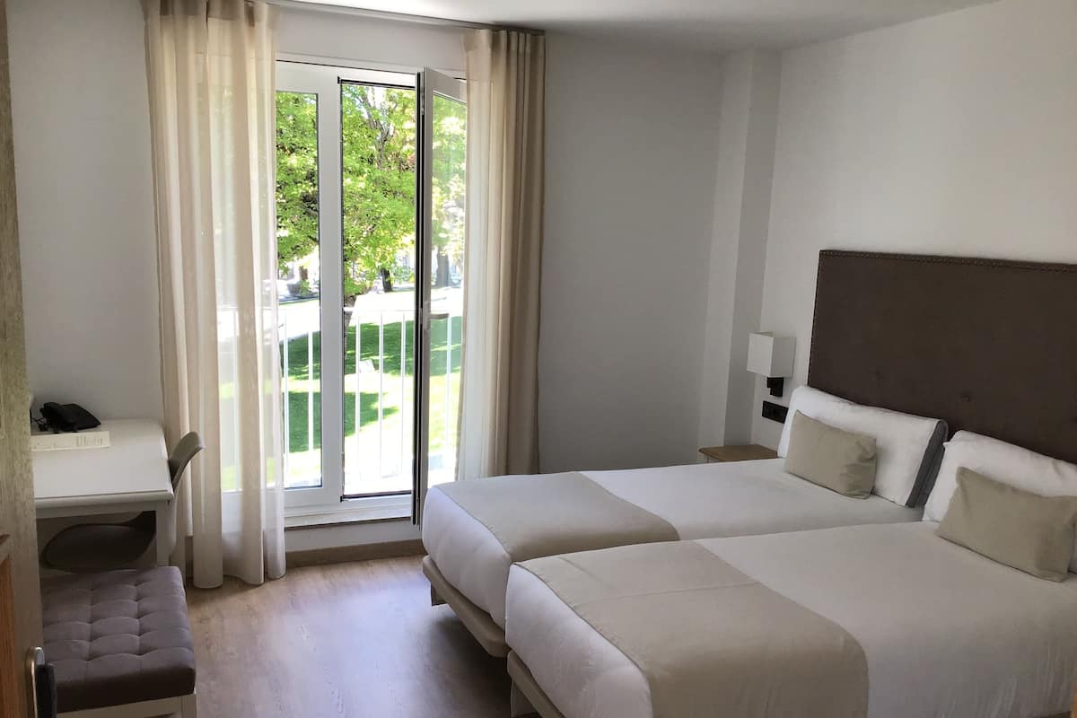 A hotel room with a bed, a desk, a chair, a lamp, and a view of the outdoors.