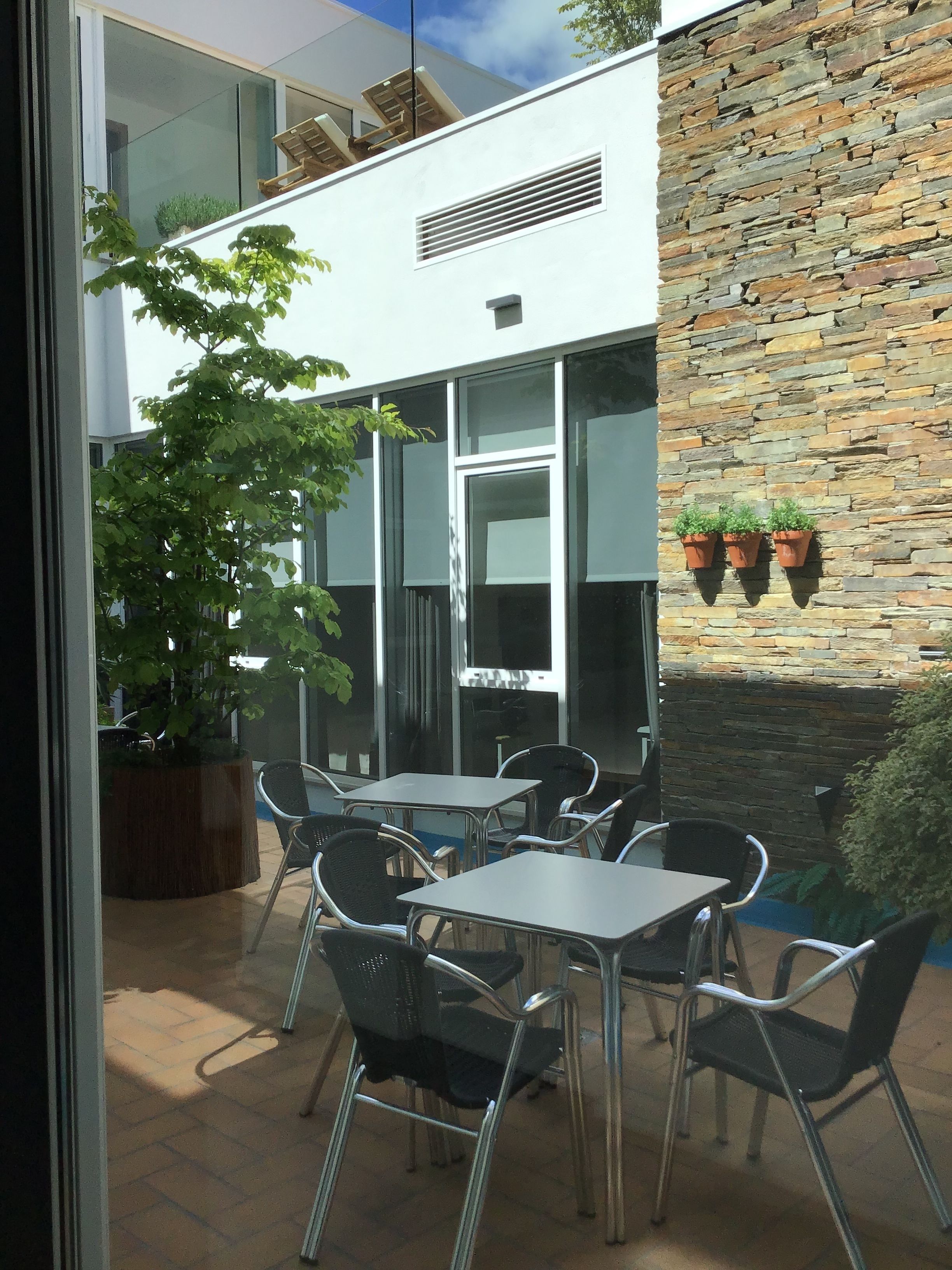 An outdoor patio with metal tables and chairs, a large potted plant, and a stone wall with hanging planters.