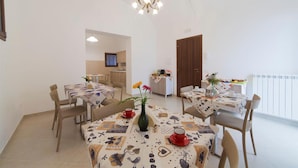 A dining area with multiple tables, each covered with a patterned tablecloth. There are chairs around the tables, and a chandelier hangs from the ceiling. A kitchen is visible in the background. - Sole di Trapani (Trapani)