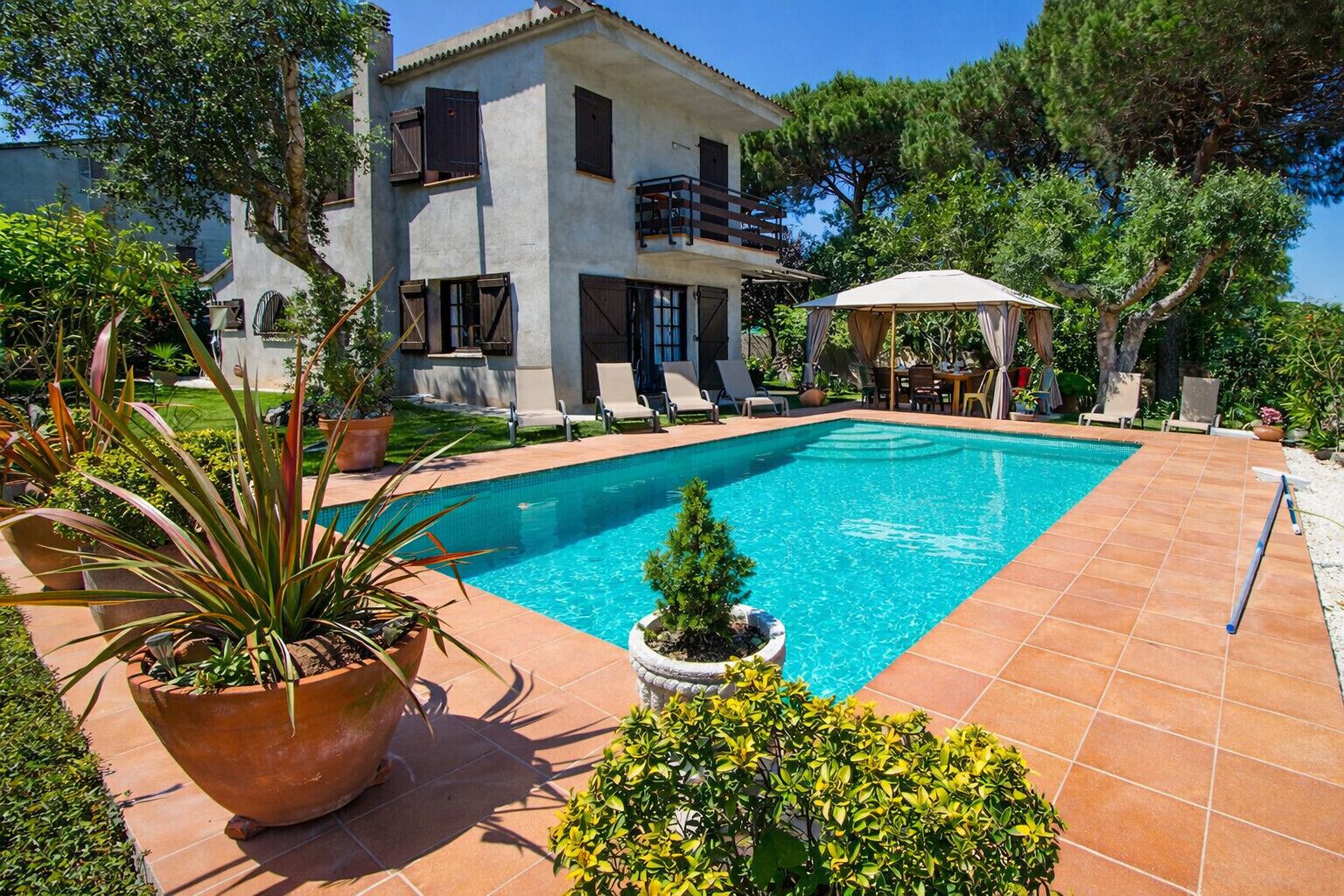 A swimming pool with a tiled deck, surrounded by potted plants and a two-story building with balconies.