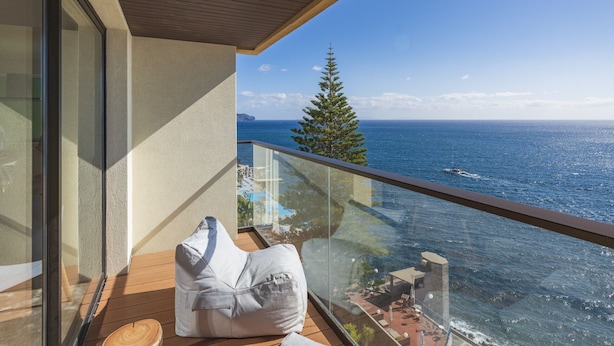A balcony with a wooden floor, a white cushioned chair, and a glass railing overlooking the sea. - Baía I, apartment with luxury design (Funchal)