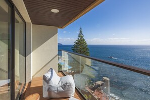 A balcony with a wooden floor, a white cushioned chair, and a glass railing overlooking the sea. - Baía I, apartment with luxury design (Funchal)