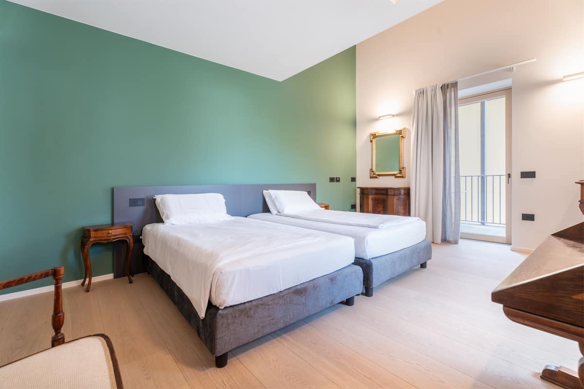 A hotel room with two beds, a wooden nightstand, a chair, a desk, a mirror, and a balcony with curtains.