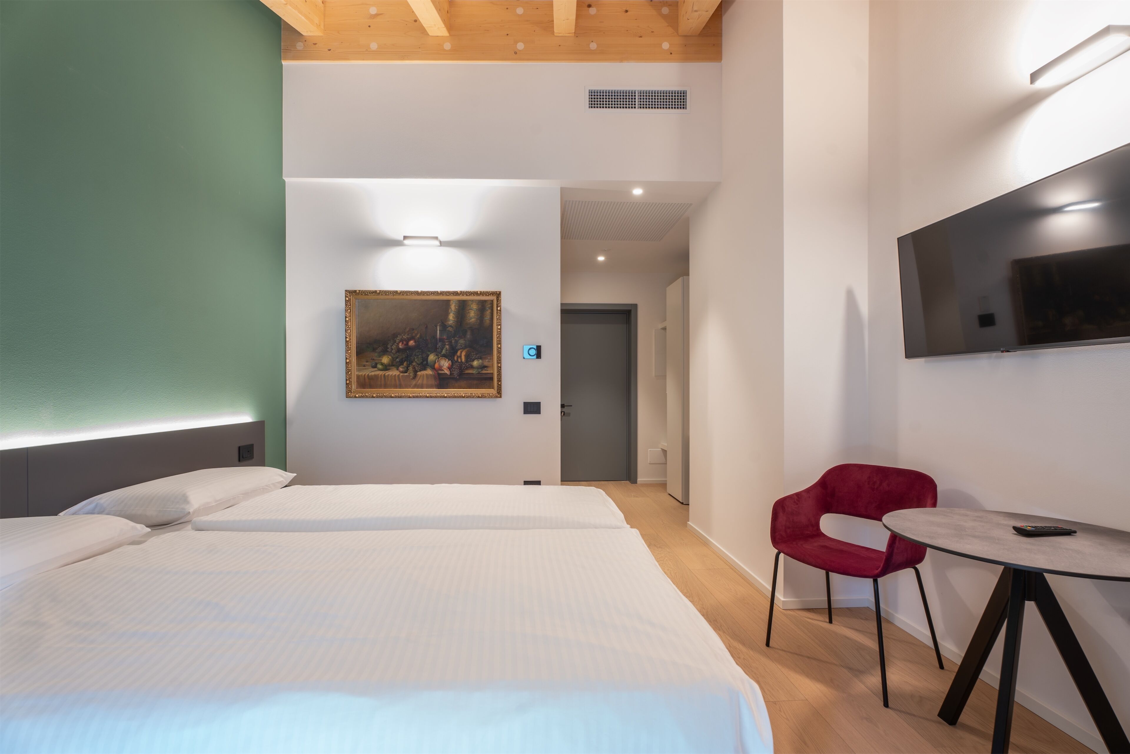A hotel room with two beds, a red chair, a round table, a painting on the wall, and a TV mounted on the wall.