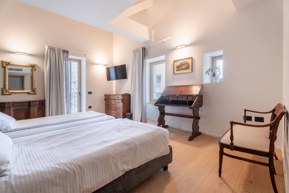 A bedroom with a large bed, a wooden dresser, a desk with a piano, a chair, and a television on the wall.