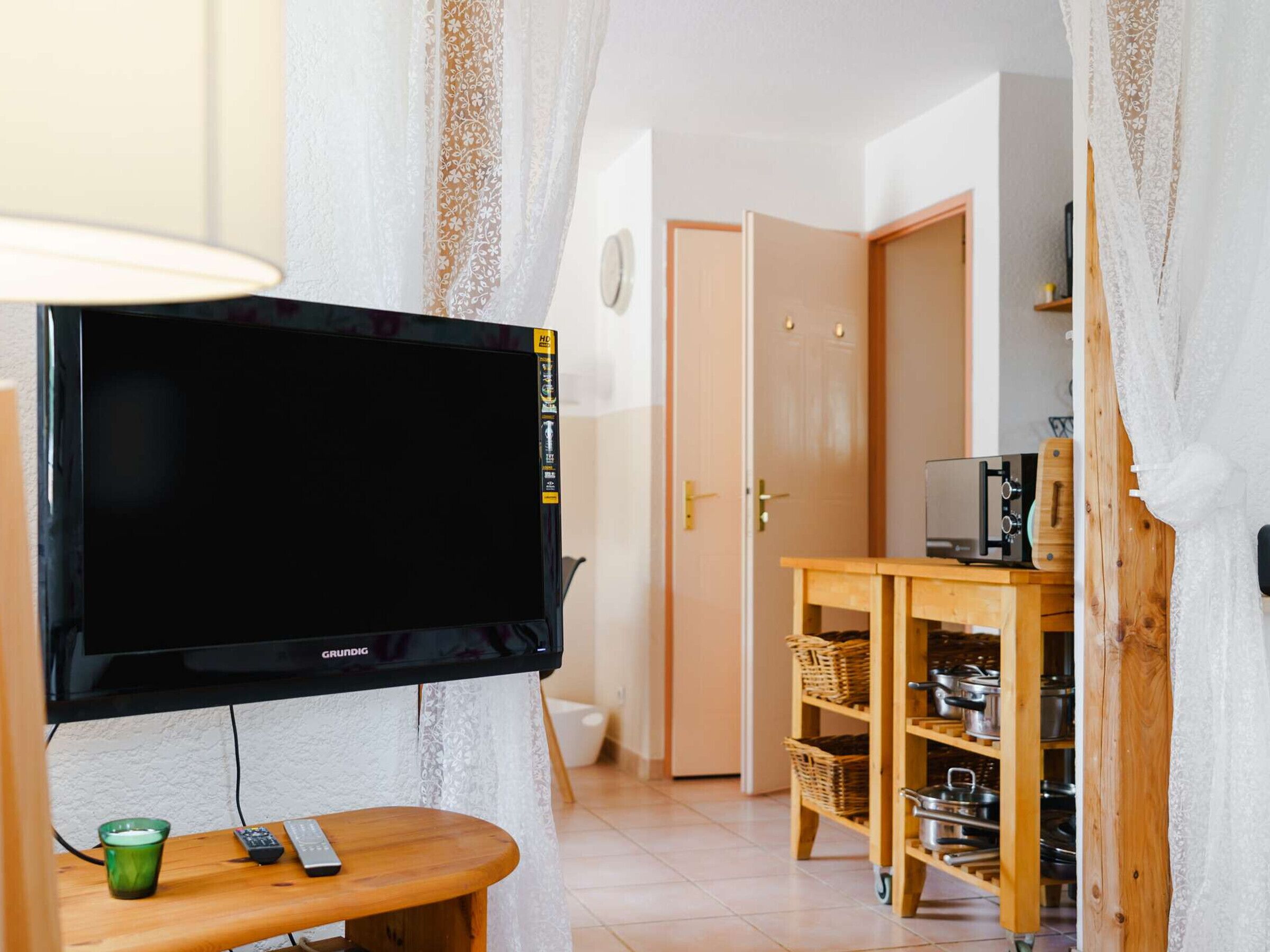Ein Wohnzimmer mit einem an der Wand befestigten Fernseher, einem Holztisch mit Fernbedienung, einem grünen Glas und einer Küchenzeile mit Regalen und Körben.