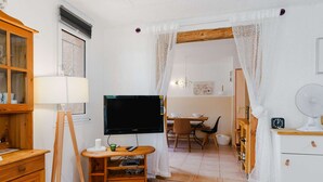 Een gezellige kamer met een televisie op een houten standaard, een eettafel met stoelen en een wit kastje.