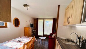Uma cozinha compacta com armários de madeira, uma área de refeições com mesa e cadeiras, uma televisão pequena e um sofá vermelho ao fundo.