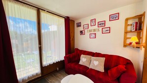 Uma sala de estar aconchegante com um sofá vermelho, duas mesinhas brancas e uma estante de madeira com livros e um abajur.