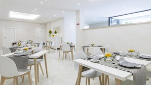 Un comedor moderno con mesas y sillas blancas, dispuesto en un espacio amplio con techo alto e iluminación empotrada.