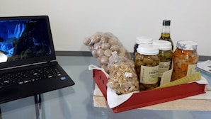Ein Laptop auf einem Tisch mit einer roten Schale, die Nüsse, Gläser mit Essiggurken und eine Flasche Öl enthält.