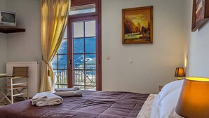 A hotel room with a bed, a desk, a chair, a radiator, a window with curtains, and a painting on the wall. - To Kallion (Karpenisi)