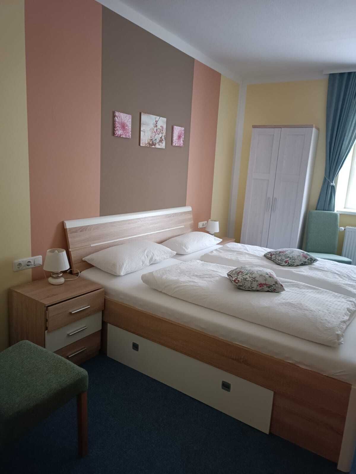 Habitación de hotel con una cama doble, una mesita de noche con lámpara, una silla pequeña y un armario.