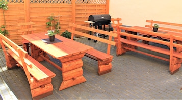 Um alpendre de madeira ao ar livre com mesa, bancos, churrasqueira e plantas em vasos.