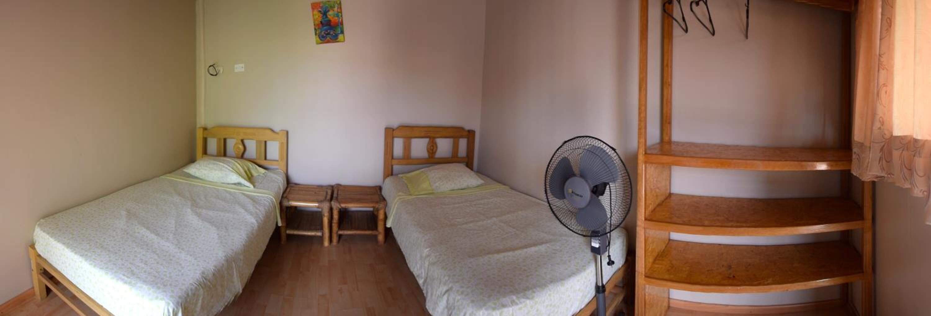 A small, simple room with two single beds, a wooden nightstand, a standing fan, and a wooden staircase.