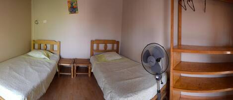 A small, simple room with two single beds, a wooden nightstand, a standing fan, and a wooden staircase.