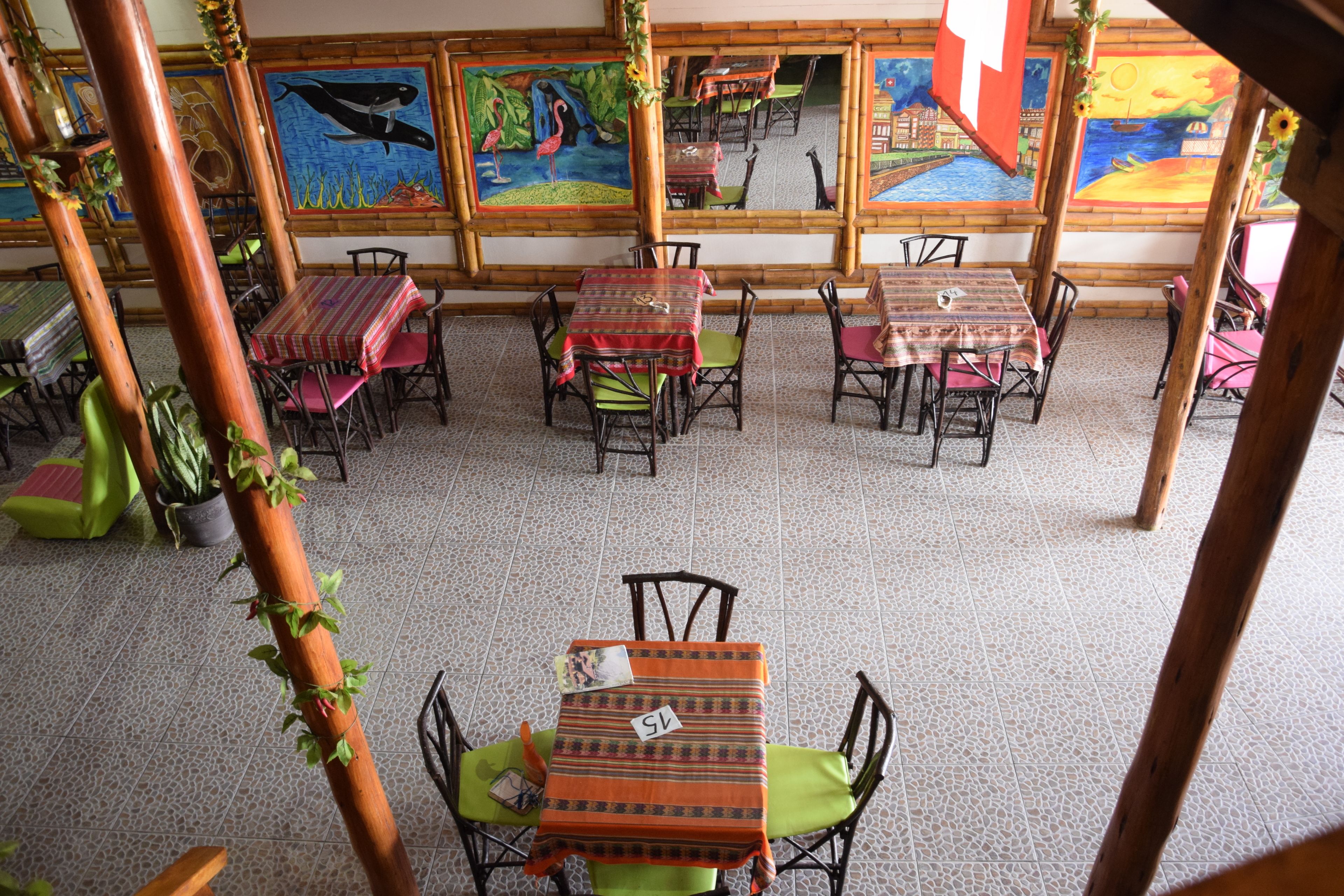 A restaurant with multiple tables and chairs, each with a tablecloth. There are paintings on the walls and a tiled floor.