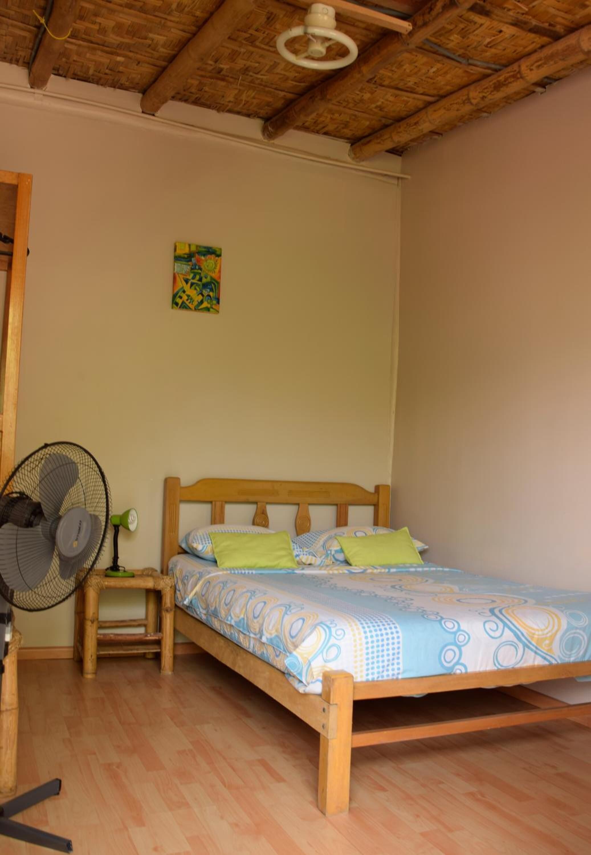 Un letto singolo con struttura in legno e biancheria a fantasia, una ventola da terra e un comodino con una lampada.