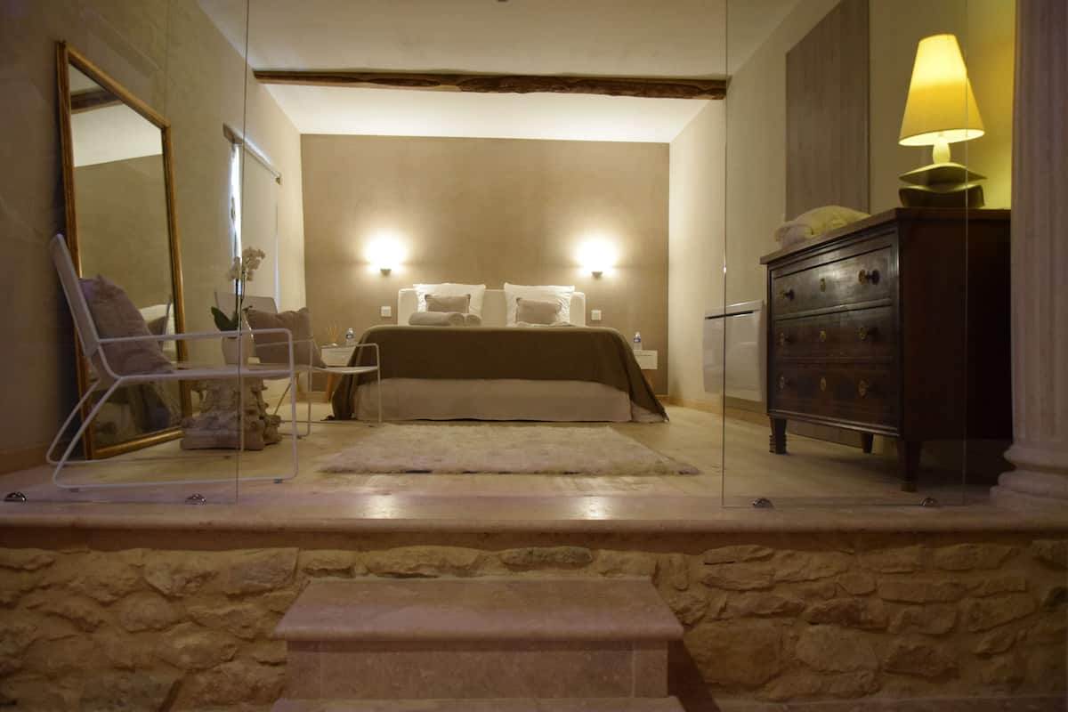 A bedroom with a stone wall, a bed with white bedding, a wooden dresser, and a mirror.
