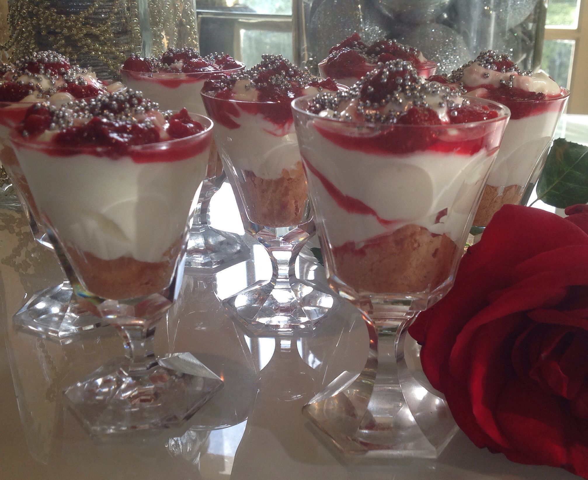 Dessert cups with layers of cream and berries, garnished with silver beads, on a reflective surface with a red rose nearby.