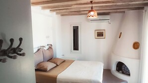 A bedroom with a bed, a small window, a wall-mounted heater, and a fireplace. - House with character (Tinos)