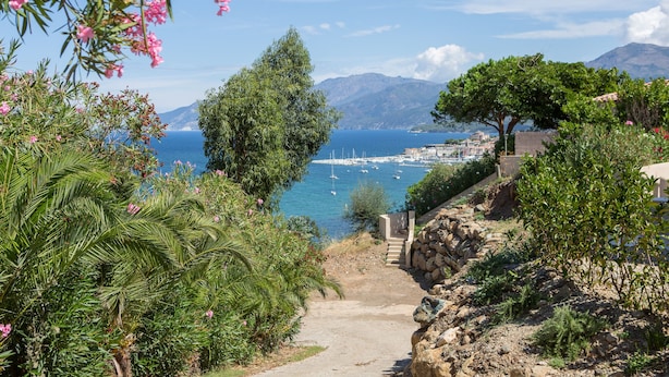 Un sentier côtier mène à une marina où sont amarrés des bateaux, le tout étant entouré d’une végétation luxuriante et avec des montagnes en toile de fond.