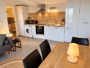 Una cuina moderna amb electrodomèstics integrats, zona de menjador amb taula i cadires, i saló amb sofà i làmpada.