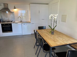 A modern kitchen with white cabinets, a stainless steel range hood, a dishwasher, and a sink. There is a wooden dining table with chairs and a potted orchid on the table.