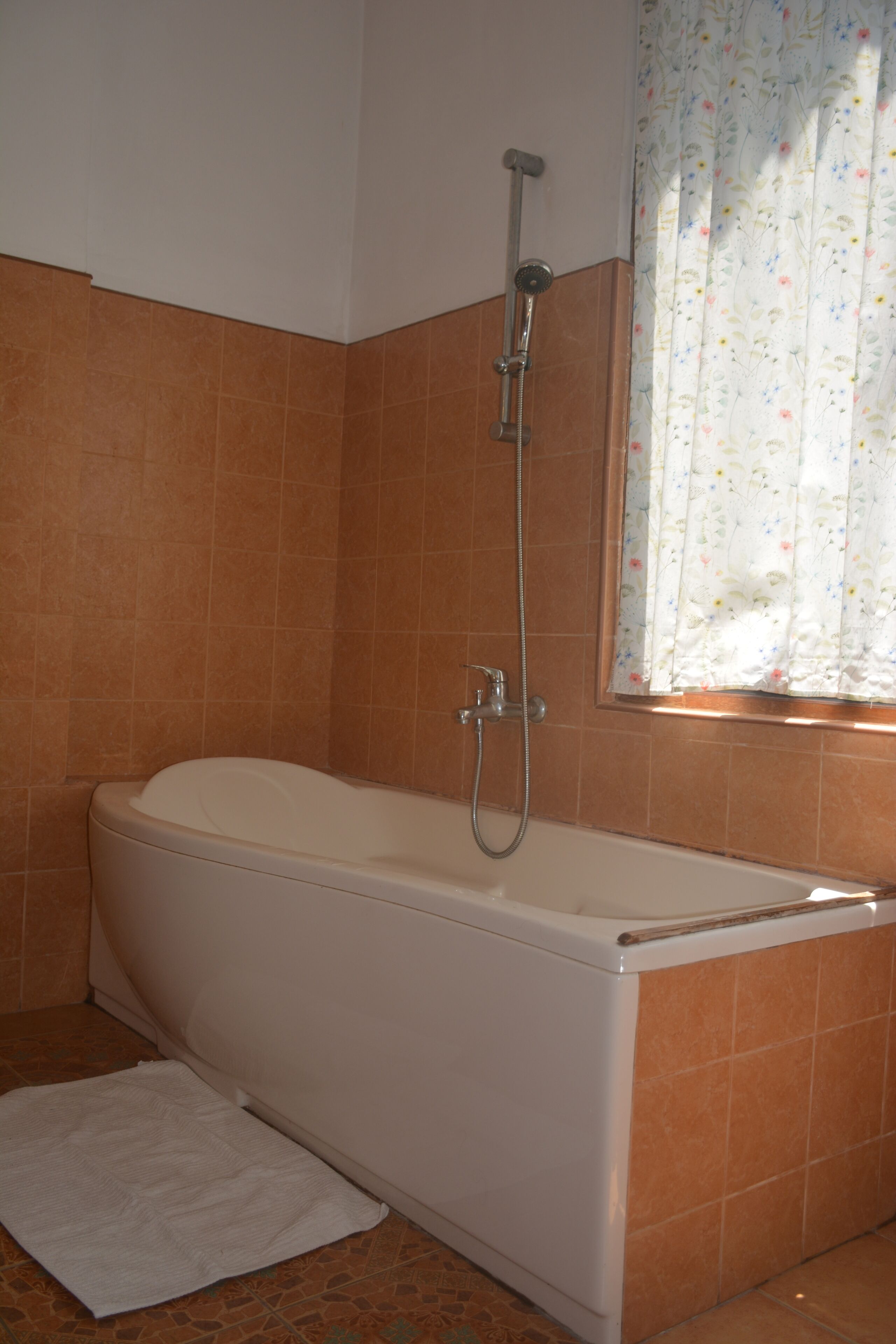 A bathroom with a bathtub, shower, and a window with floral curtains.