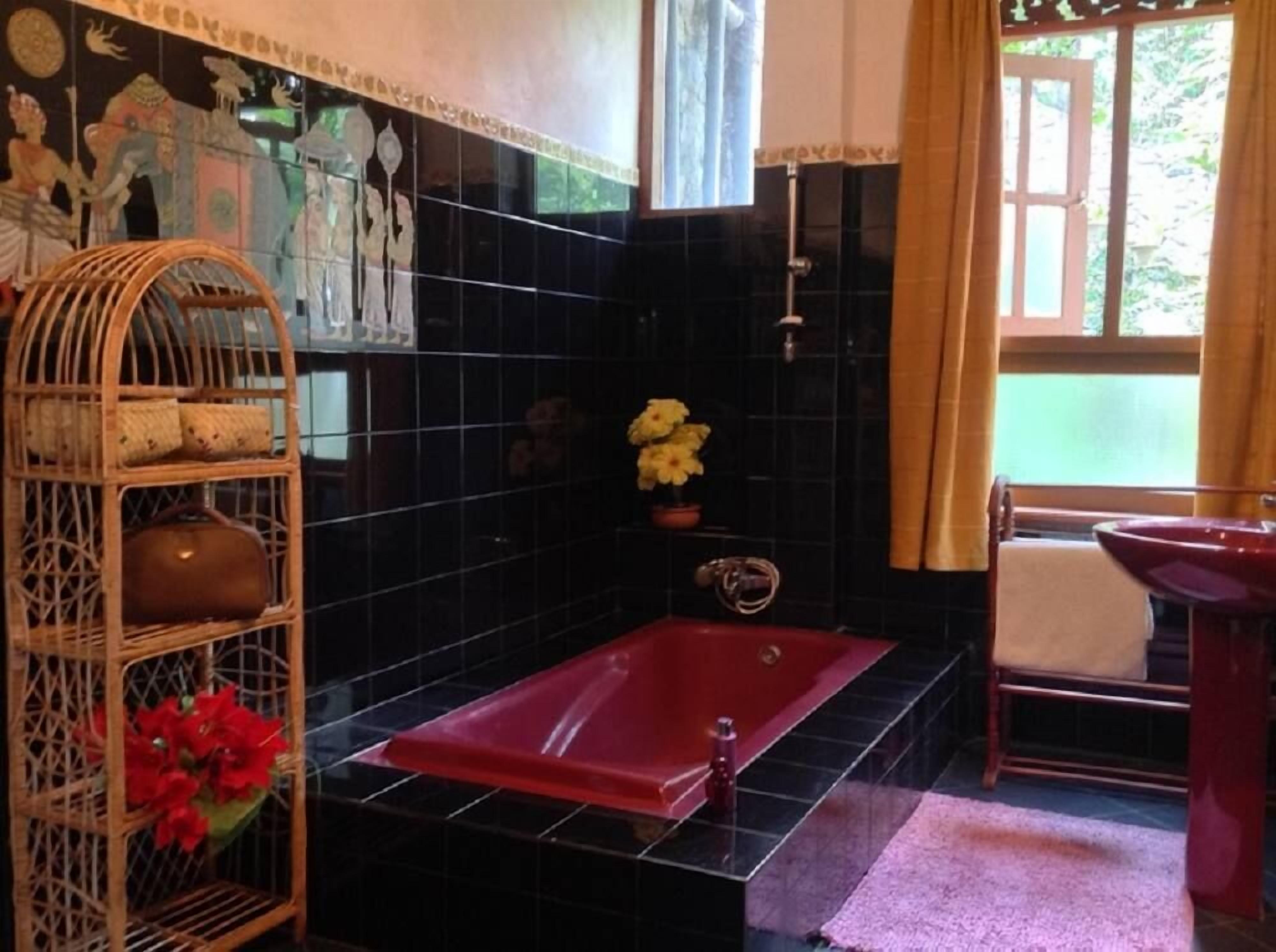 A bathroom with a red bathtub, black tiled walls, a wicker shelf with toiletries, a potted plant, and a window with curtains.