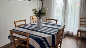 Un coin repas avec une table recouverte d’une nappe à rayures, entourée de chaises en bois. Un vase rempli de fleurs est posé sur la table. La pièce est dotée de poutres apparentes en bois au plafond et d’une fenêtre avec des rideaux.
