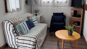 Un salon chaleureux avec un canapé à rayures, un fauteuil sombre, une table basse en bois ornée d’une plante et un téléviseur posé sur une étagère en bois.