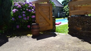 A wooden gate leading to a stone wall, a barrel, and purple flowers.