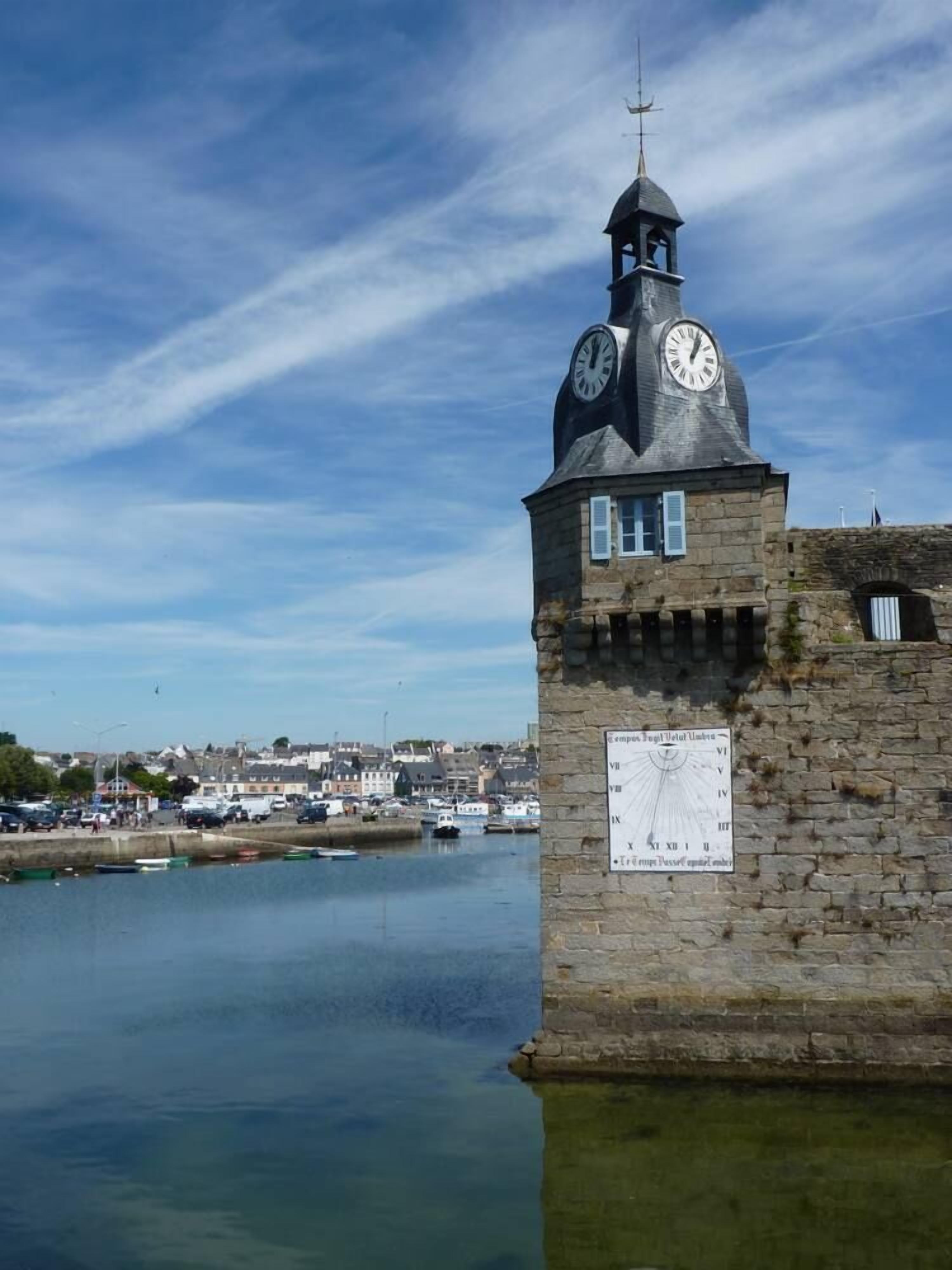 A stone clock tower with a weather vane, located near a body of water with boats and buildings in the background.