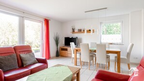 Un salon moderne avec un canapé rouge, une table à manger avec des chaises, un téléviseur posé sur un meuble en bois et de grandes fenêtres avec des rideaux.