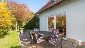 Une terrasse avec une table et des chaises, un jardin et une maison avec un toit en tuiles.