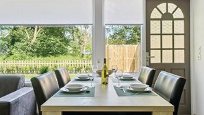 Un coin repas avec une table dressée pour quatre personnes, une bouteille de vin et une vue sur un jardin avec une clôture en bois et de la verdure.