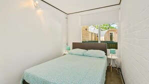 Une petite chambre à coucher moderne, avec un lit simple, une table de chevet, un miroir et une fenêtre donnant sur une structure en bois à l’extérieur.